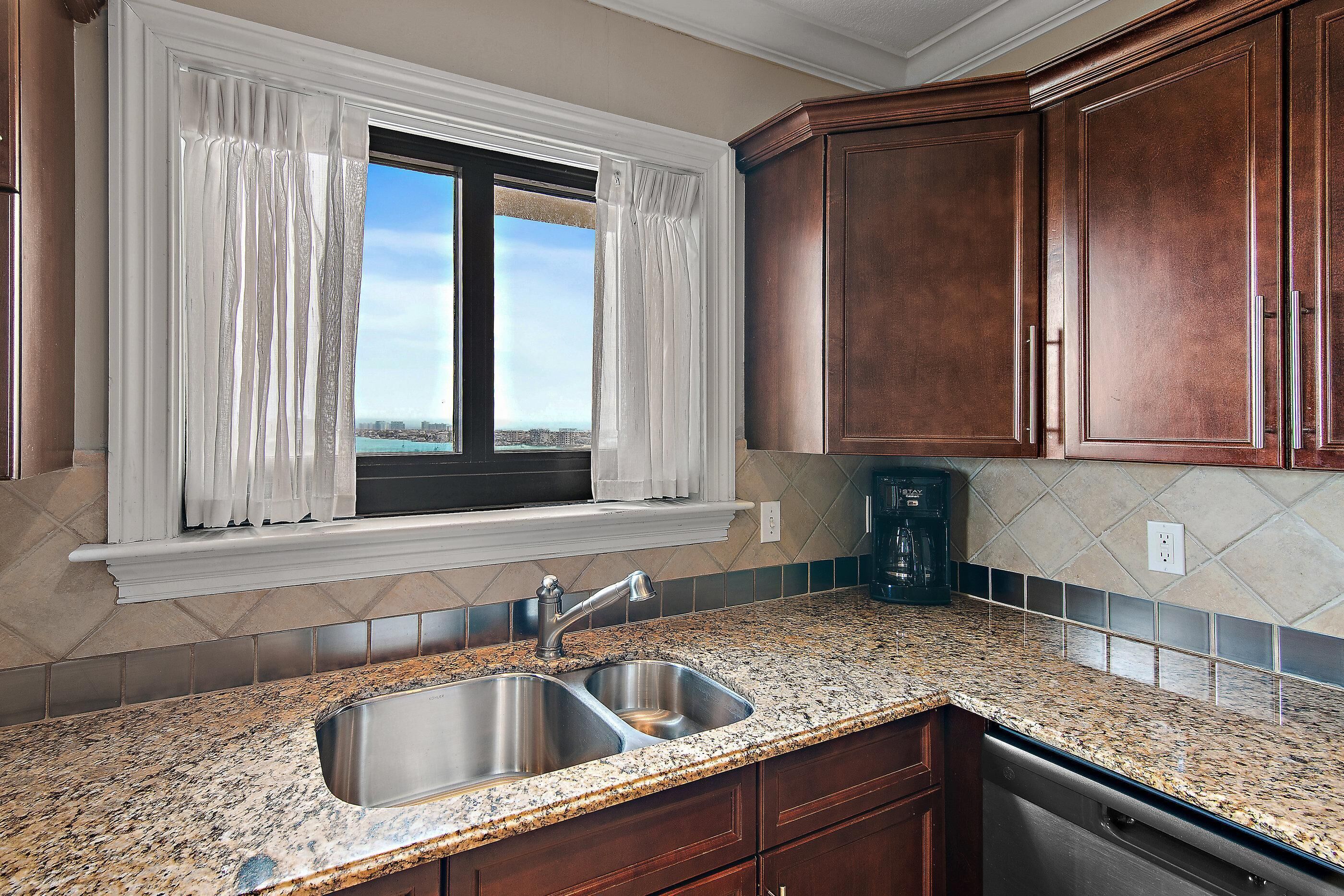 A kitchen with granite countertops, stainless steel sink, wooden cabinets, and a window with white curtains.