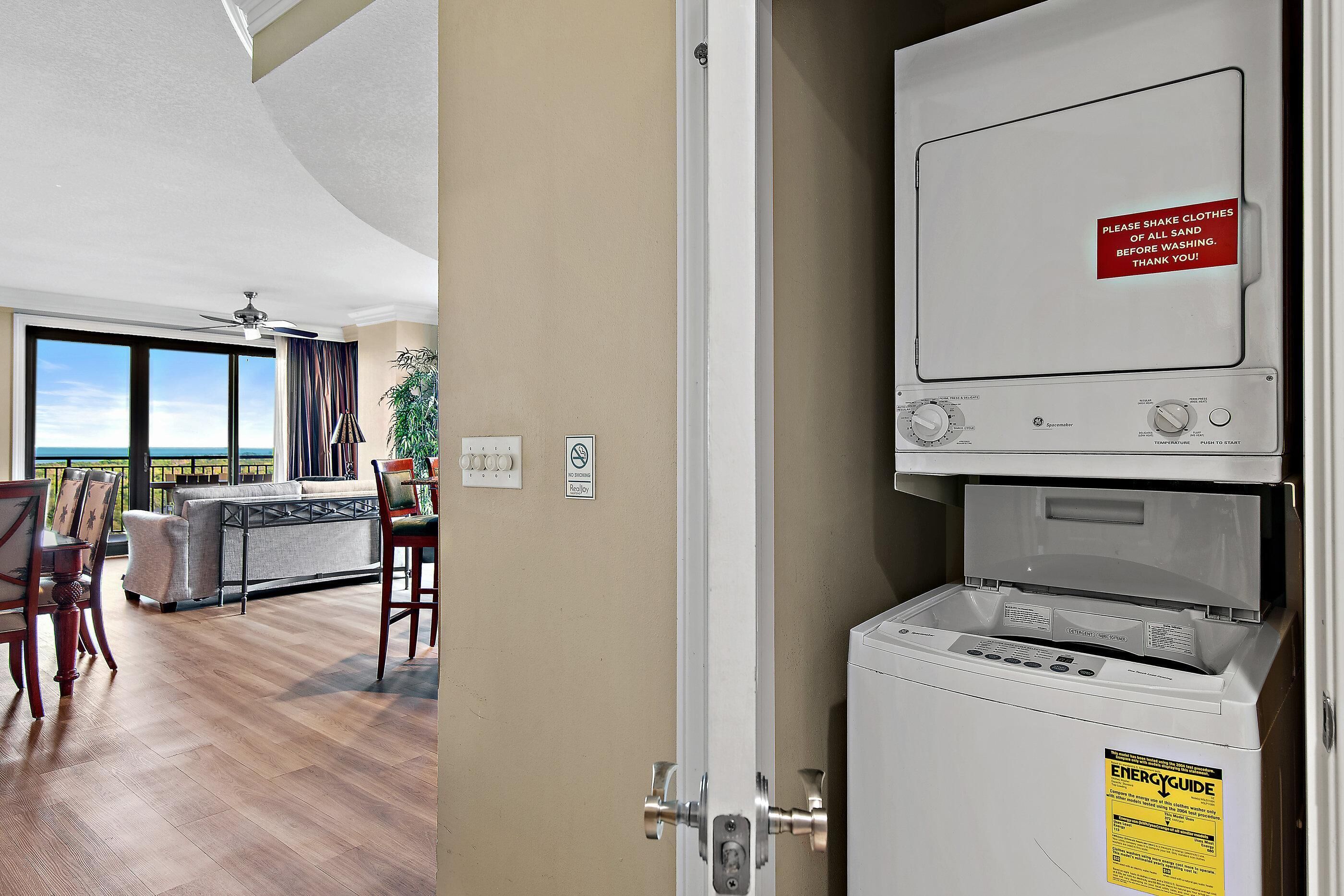A laundry room with a GE EnergyGuide label, a sign instructing to shake clothes before washing, and a view of a living area with a sofa, chairs, and a plant.