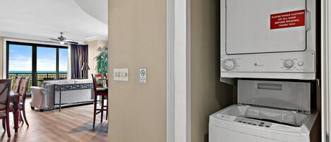 A laundry room with a GE EnergyGuide label, a sign instructing to shake clothes before washing, and a view of a living area with a sofa, chairs, and a plant.