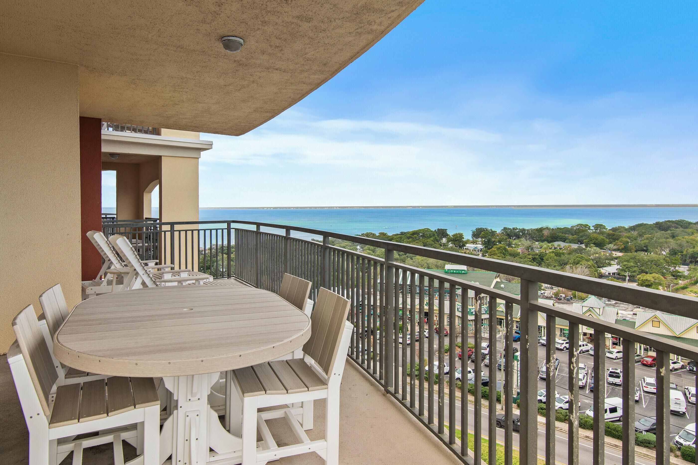 A balcony with a round table and chairs, overlooking a parking area and a view of the sea.