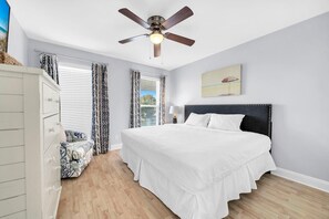 Quarto com cama grande, ventilador de teto, mesa de cabeceira, cômoda e janela com cortinas.