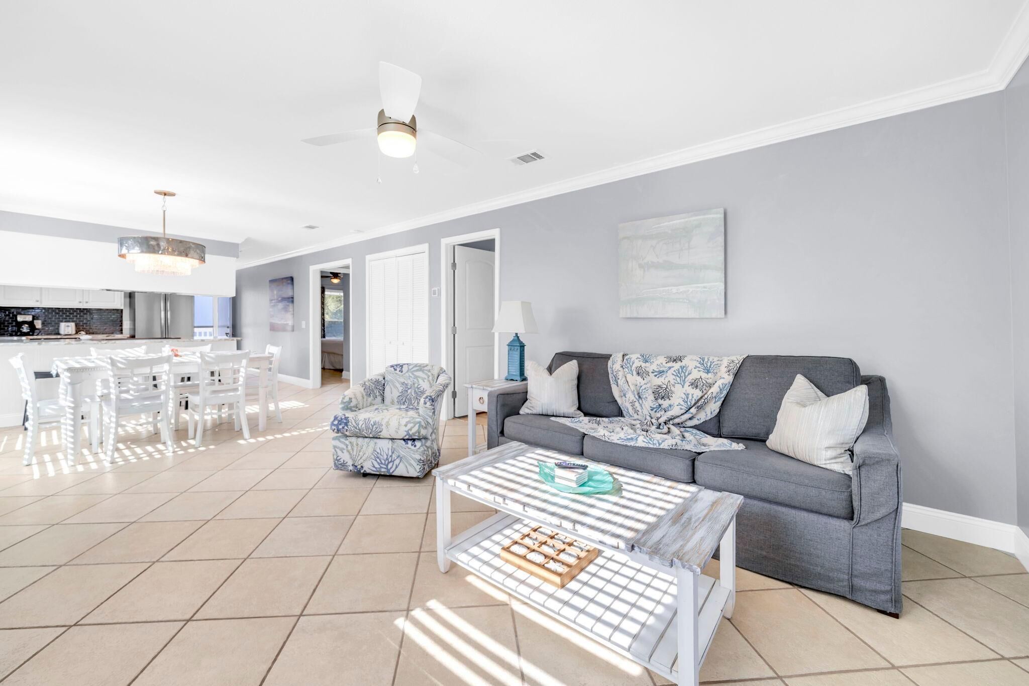 A spacious living room with a grey sofa, a coffee table, and a patterned armchair. There is a dining area with white chairs and a chandelier.