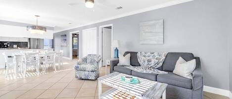 A spacious living room with a grey sofa, a coffee table, and a patterned armchair. There is a dining area with white chairs and a chandelier.