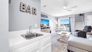 A modern kitchen with a sink and cabinets, a dining area with a table and chairs, and a living area with a sofa and a chair. - Lullwater Beach 415 (Panama City Beach)