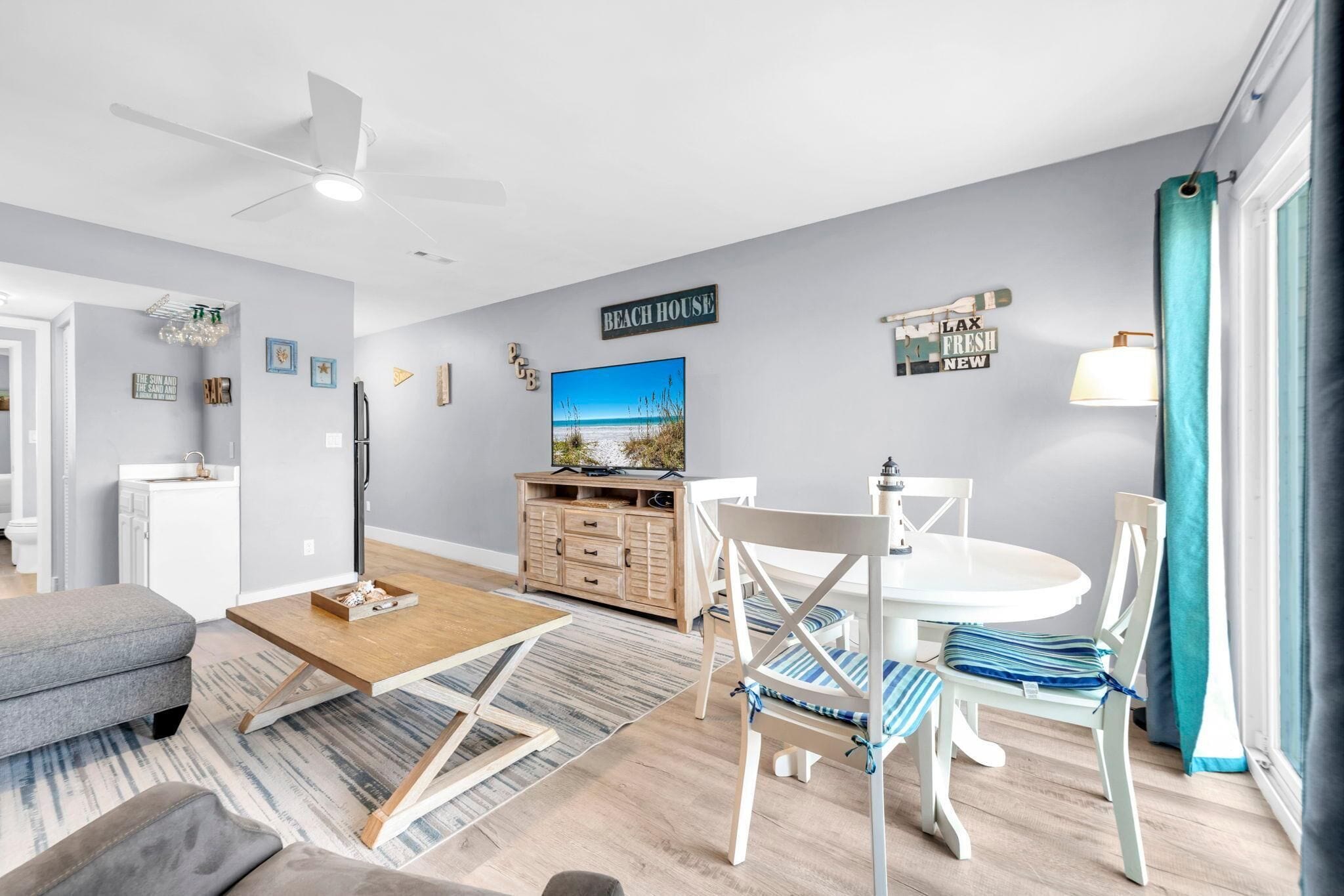 A modern living room with a dining area, a ceiling fan, a painting of a beach, and a sign that reads "BEACH HOUSE".
