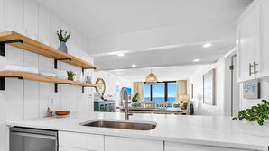 A modern kitchen with white cabinetry, a stainless steel sink, and a view to the living area. - Edgewater Tower 3-1003 (Panama City Beach)