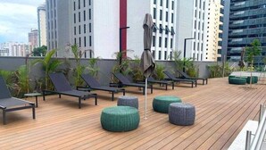 A rooftop deck with lounge chairs and ottomans, surrounded by tall buildings. - Studios with outdoor pool in the lounge. (São Paulo)