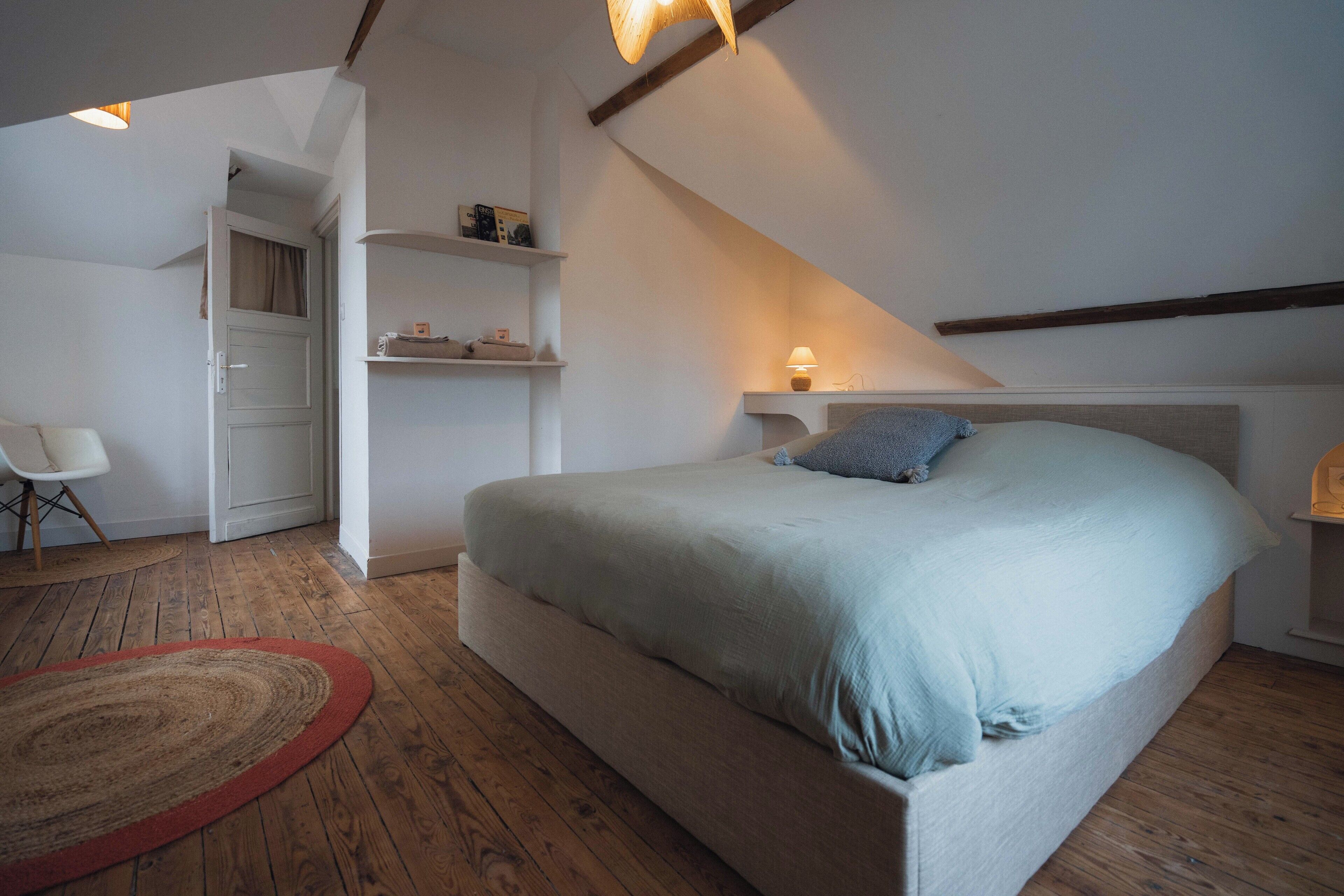 A bedroom with a bed, wooden flooring, a red rug, a chair, and a shelf with items.