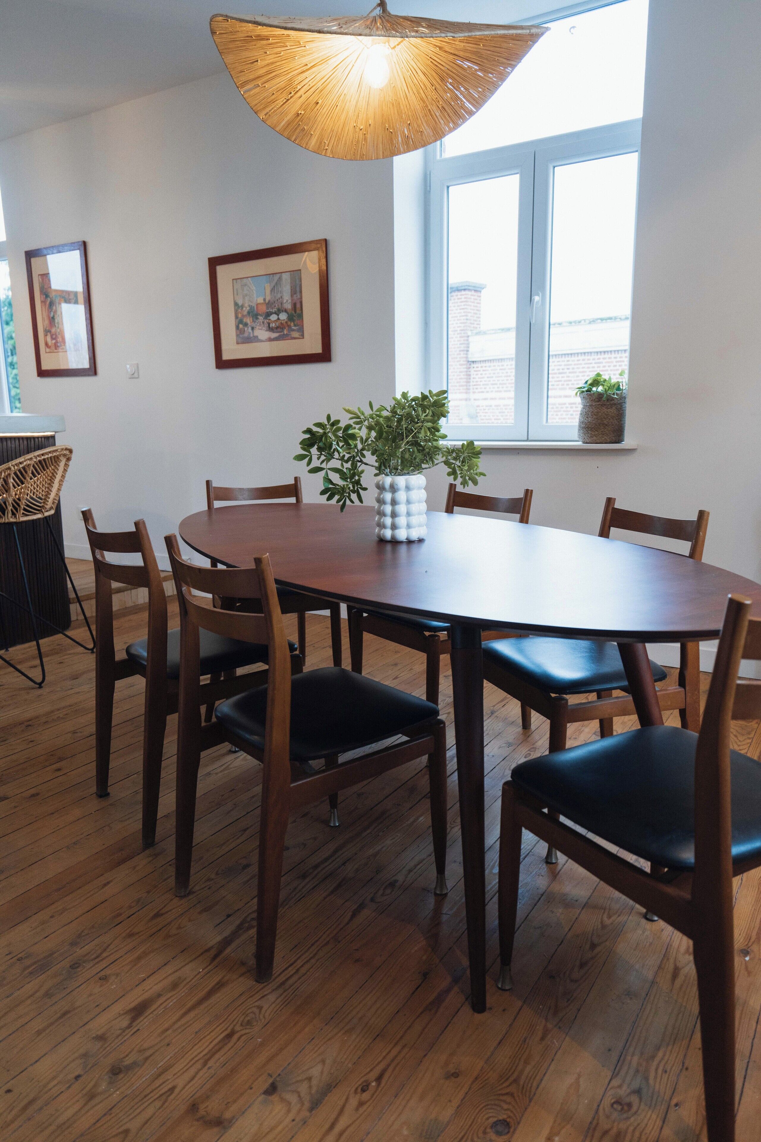 Un comedor con una mesa redonda de madera, seis sillas, una lámpara colgante y una ventana con vistas a edificios.