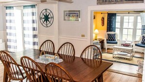 Un comedor con mesa de madera y seis sillas, decoración de pared con brújula y vista a una playa a través de un ventanal abierto.