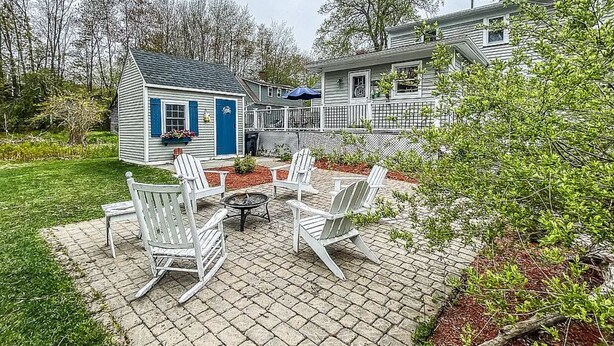 Un patio con sillas Adirondack blancas, un pequeño hogar para fogata y un cobertizo con puerta azul.