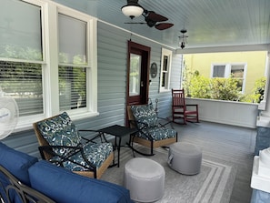 A covered porch with wicker furniture, a fan, and a view of a yellow building.