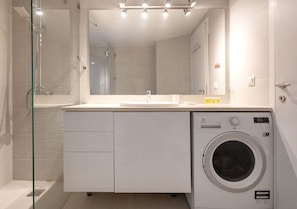 A modern bathroom with a glass shower enclosure, a built-in sink, a mirror, and a front-loading washing machine. - Private Garden Duplex in Voula by GHH (Voula)