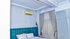 A hotel room with a bed, white bedding, a teal headboard, an air conditioning unit, a wooden nightstand, and a large mirror. - The Precision (Lagos)