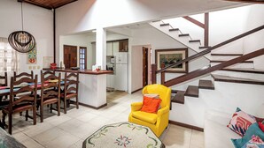 A spacious living area with a wooden ceiling, a dining table with chairs, a yellow armchair, a staircase, and a kitchen in the background. - Cozy house with pool in the center of Buzios. (Buzios)