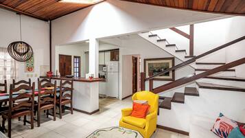 A spacious living area with a wooden ceiling, a dining table with chairs, a yellow armchair, a staircase, and a kitchen in the background.
