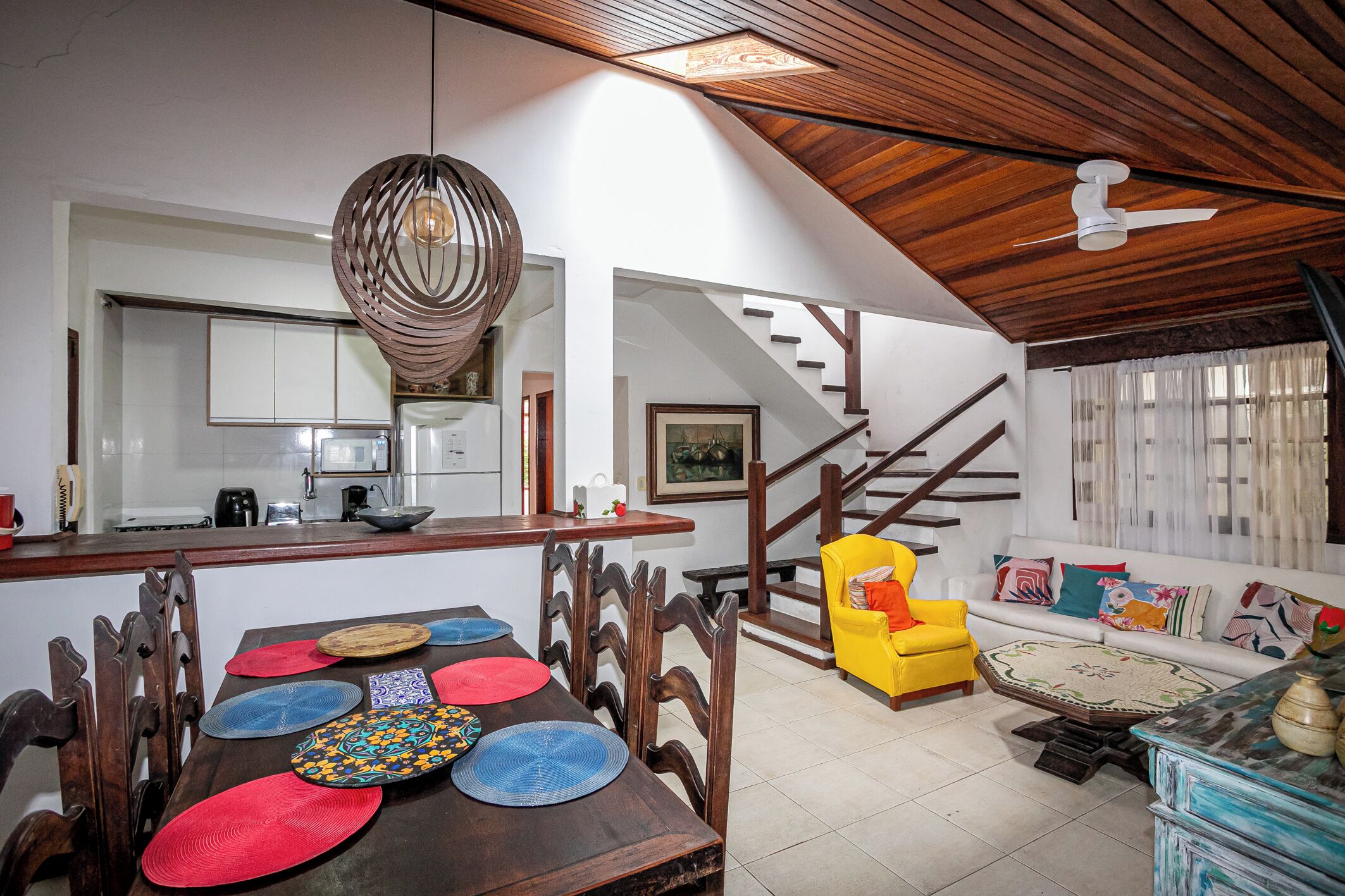 A spacious living area with a dining table set with colorful placemats, a yellow armchair, and a sofa with cushions. A staircase leads to an upper level.