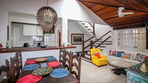 A spacious living area with a dining table set with colorful placemats, a yellow armchair, and a sofa with cushions. A staircase leads to an upper level. - Cozy house with pool in the center of Buzios. (Buzios)