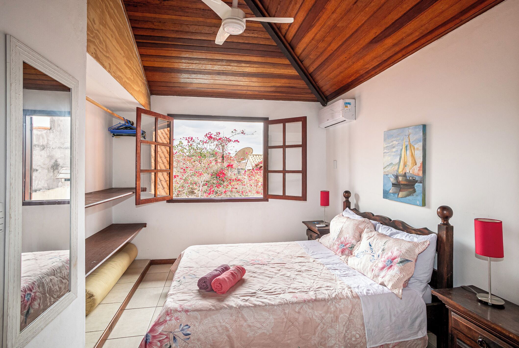 Un dormitorio con techo de madera, una cama con colcha floral, un ventanal con vista a flores rosadas, un cuadro de un velero y una lámpara roja sobre una mesita de noche.