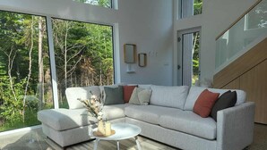 A modern living room with a large sectional sofa, a round coffee table, and a view of a forest through large windows. - Chalet Héméra - Bord de l`eau avec SPA et Sauna (Notre-Dame-de-Montauban)