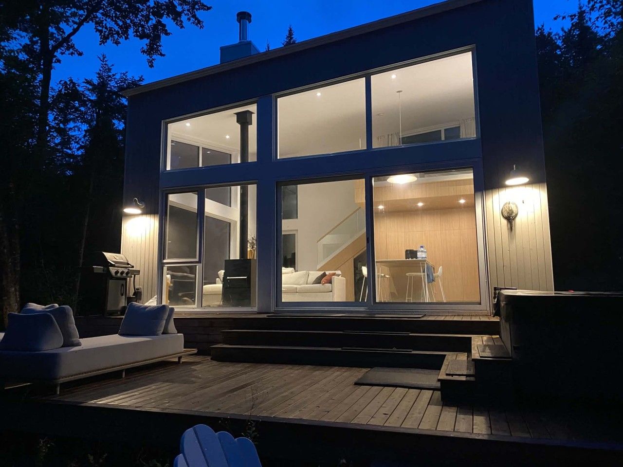 Ein modernes Haus mit großen Glasfenstern, einer Terrasse mit Sitzgelegenheiten und einem sichtbaren Innenbereich mit Sofa und Essbereich.