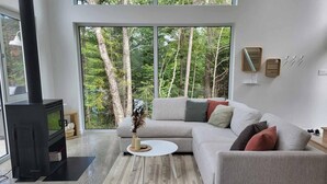 A modern living room with a large window, a fireplace, a grey sofa, and a coffee table. - Chalet Héméra - Bord de l`eau avec SPA et Sauna (Notre-Dame-de-Montauban)