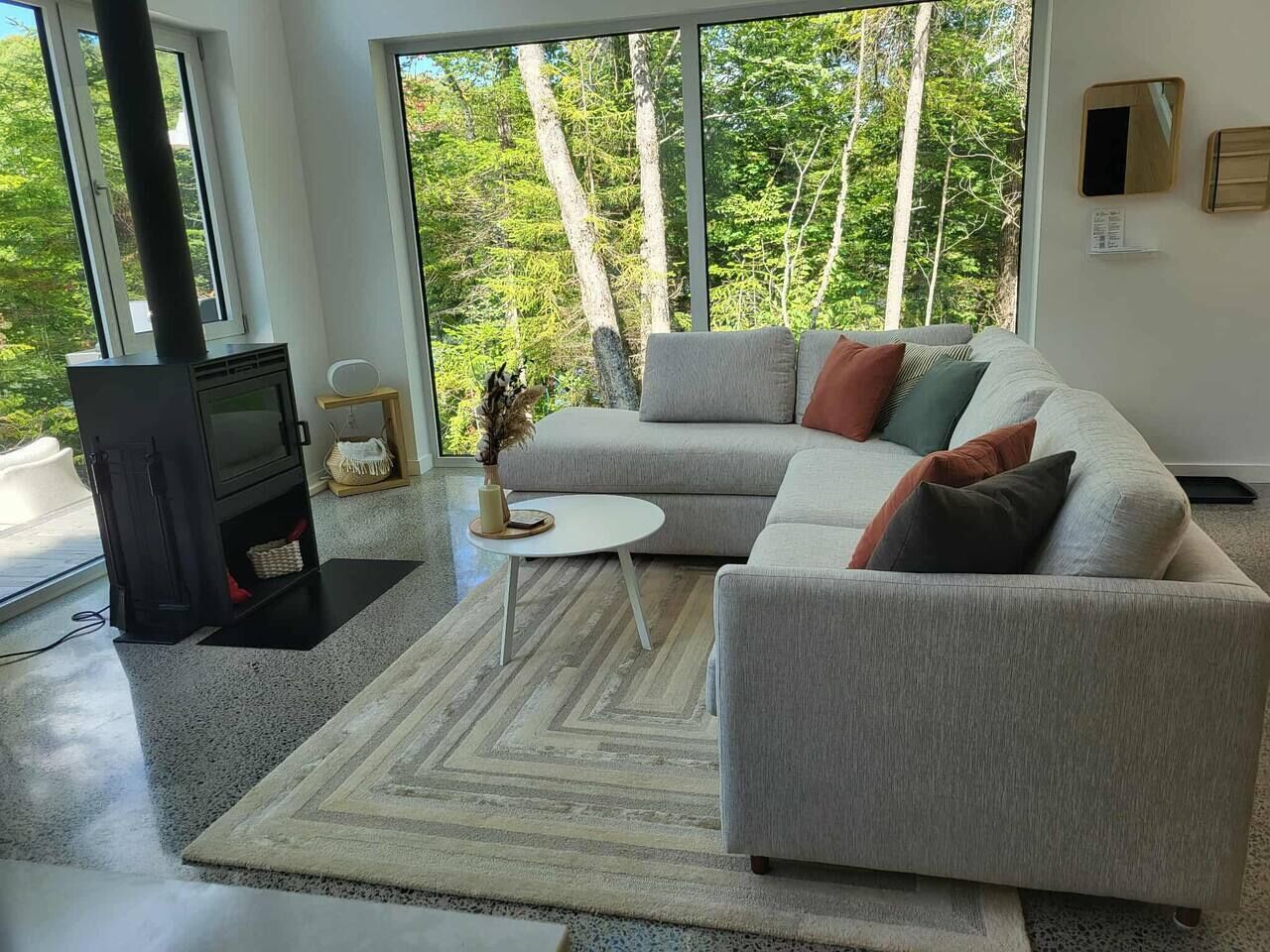 Ein modernes Wohnzimmer mit einem großen grauen Ecksofa, einem schwarzen Kamin und Blick auf einen Wald durch große Fenster.