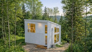 A small, single-story cabin with large windows, surrounded by dense forest. - Chalet Héméra - Bord de l`eau avec SPA et Sauna (Notre-Dame-de-Montauban)
