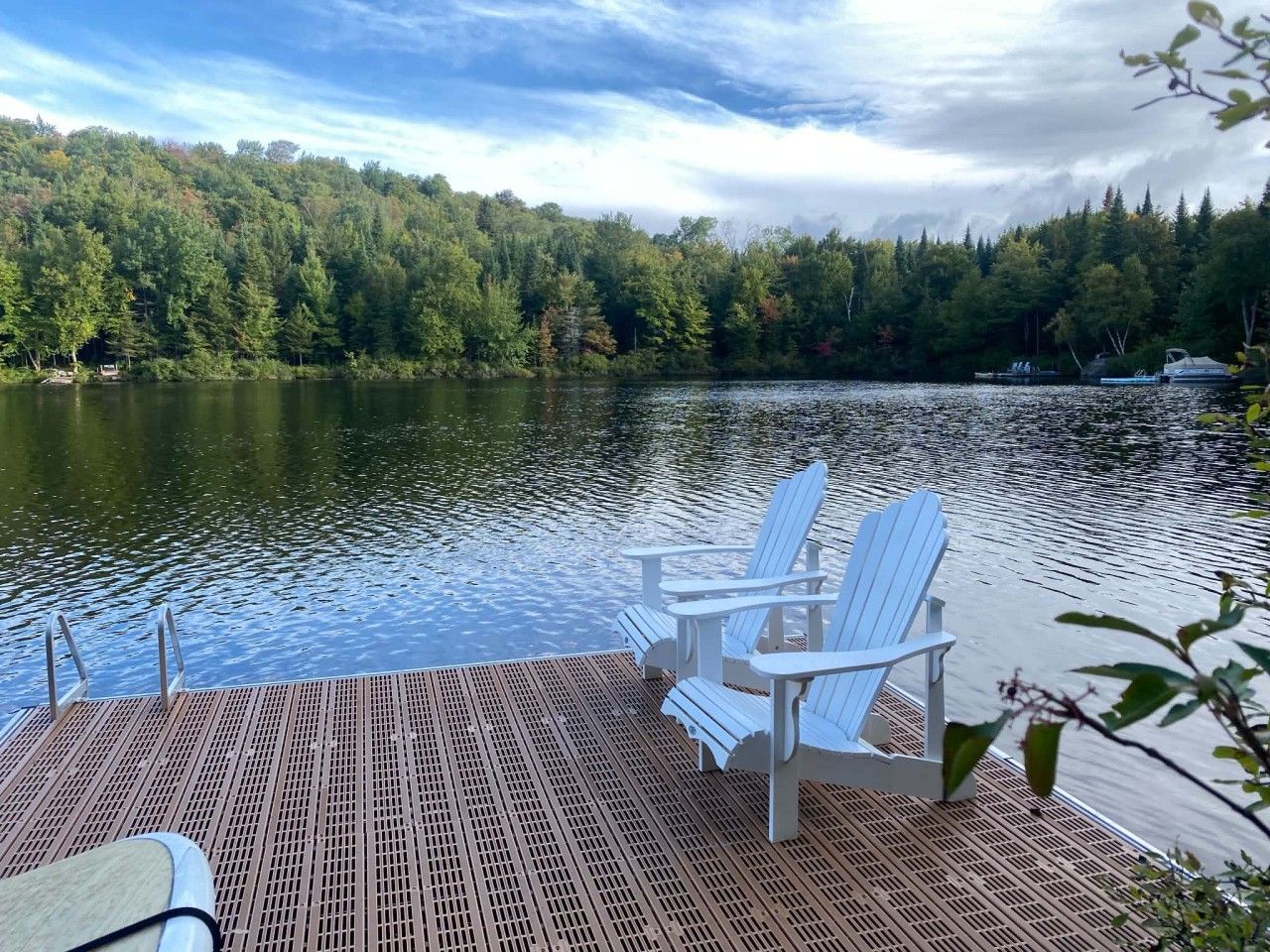 A wooden dock with two white Adirondack chairs facing a calm lake, surrounded by dense forest.