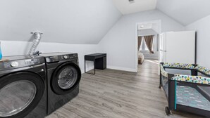 A laundry room with a washing machine and dryer, a small table, and a bed with a colorful headboard. - Modern 5BR Duplex w/ Easy NYC Access & Parking (Bayonne)