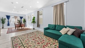 A spacious living room with a green sofa, a dining area with a table and chairs, and a large window with curtains. - Modern 5BR Duplex w/ Easy NYC Access & Parking (Bayonne)