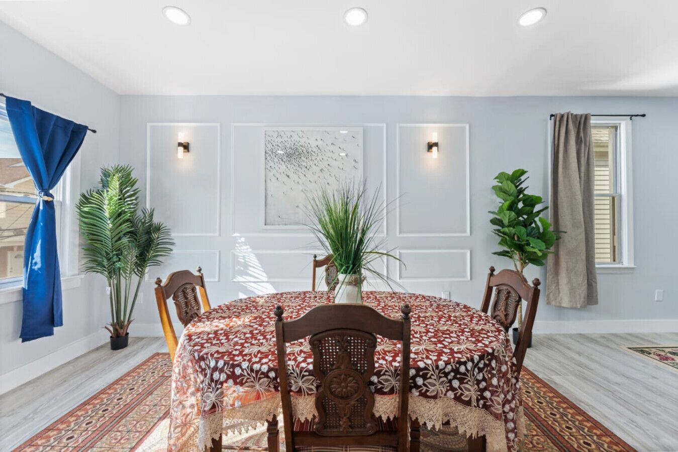 A dining room with a round table, chairs, a patterned tablecloth, potted plants, and a large abstract painting on the wall.