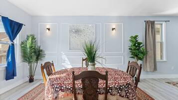 A dining room with a round table, chairs, a patterned tablecloth, potted plants, and a large abstract painting on the wall.