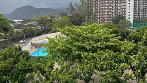 A parking area with several cars, a building with balconies, and a mountainous background. - Apto com Piscina e Sauna Perto da Praia da Barra (Rio de Janeiro)