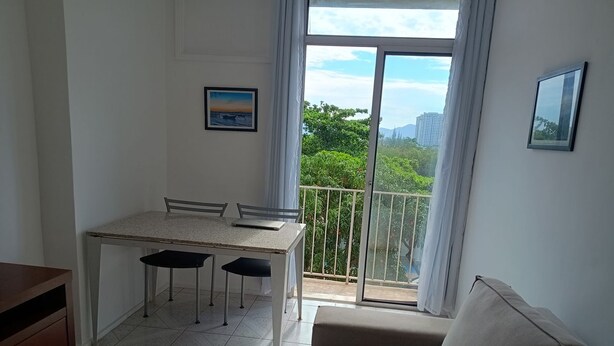 A small, bright room with a dining table and chairs, a sofa, and a balcony with a view of greenery and buildings. - Apto com Piscina e Sauna Perto da Praia da Barra (Rio de Janeiro)