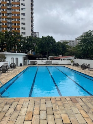 A swimming pool with clear blue water, surrounded by stone tiles and outdoor furniture. - Apto com Piscina e Sauna Perto da Praia da Barra (Rio de Janeiro)