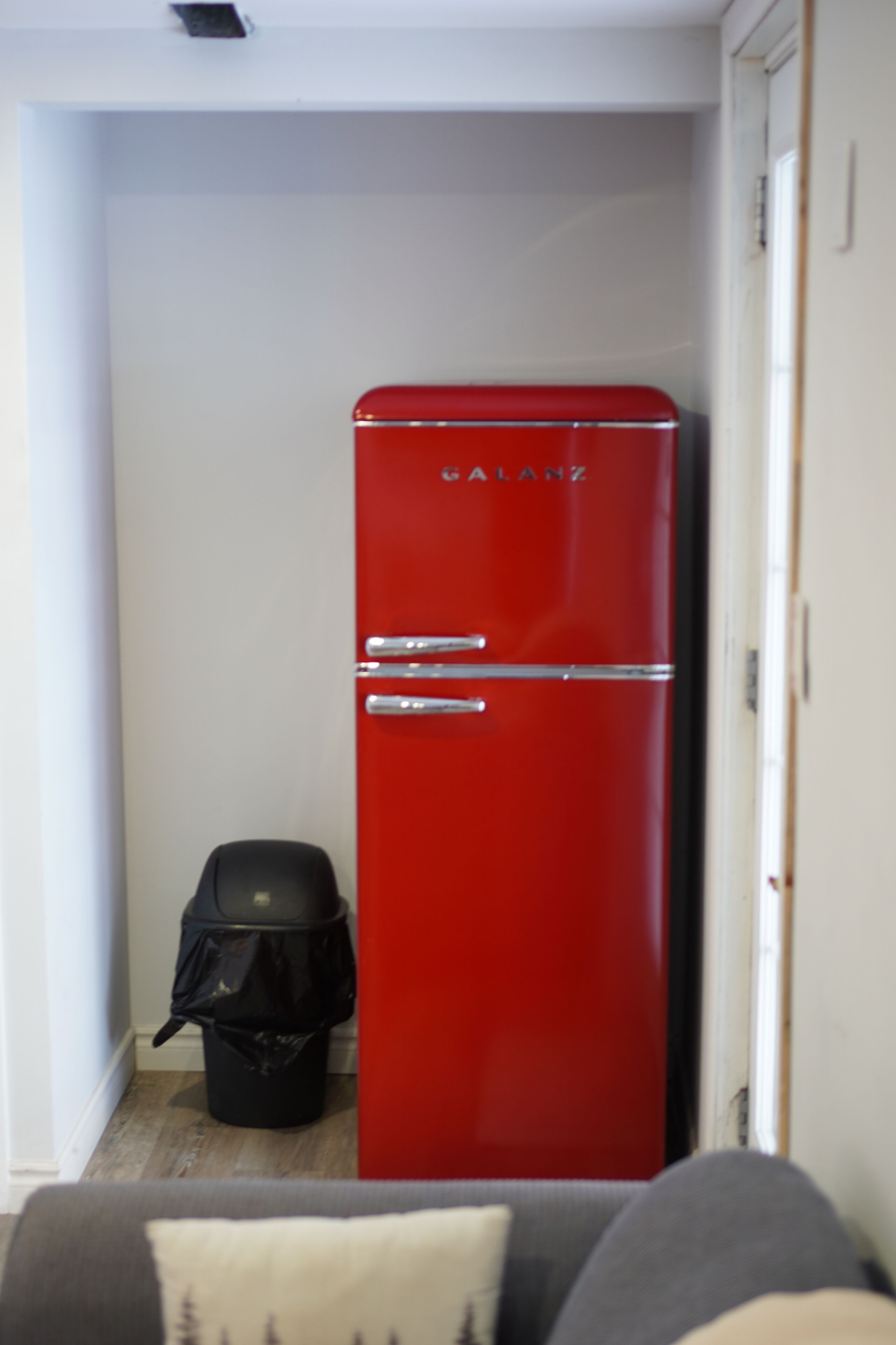 Una habitación compacta con un refrigerador vintage rojo, un bote de basura negro y un sofá gris con un cojín.