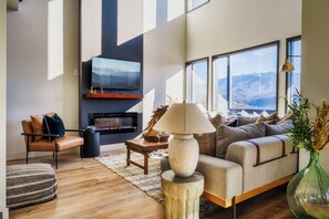 A modern living room with a sofa, a coffee table, a TV mounted on the wall, and a large window with a mountain view. - Luxury Gatlinburg Cabin w/ Indoor Pool & Mountain Views - Minutes from PG (Gatlinburg)