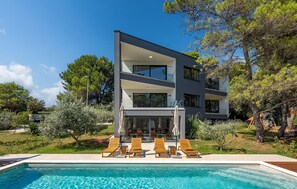 Une maison moderne à deux étages, dotée d’une piscine, d’un espace extérieur aménagé et d’une végétation luxuriante.