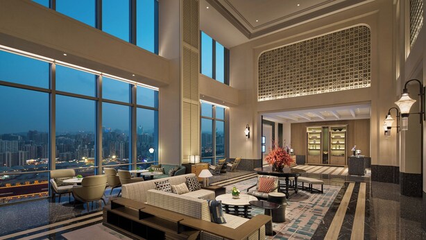 Un vestíbulo amplio con techos altos, grandes ventanales que ofrecen vistas de la ciudad, un área de descanso con sofás y sillones, y una pared decorativa con diseños intrincados.