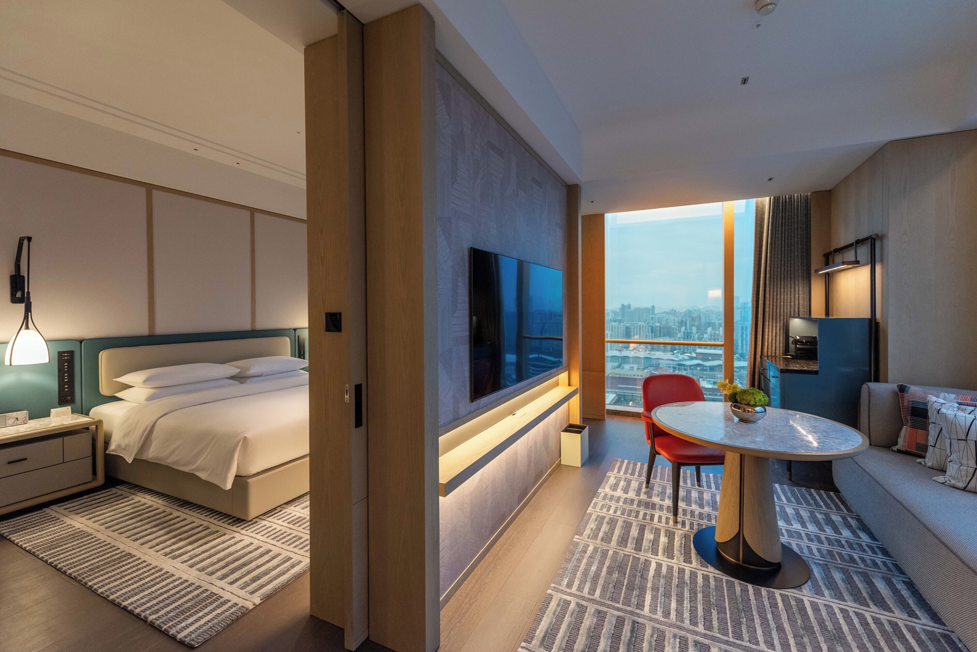 Una habitación de hotel moderna con una cama grande, un televisor, un área de comedor con mesa y sillas, y vistas a la ciudad.
