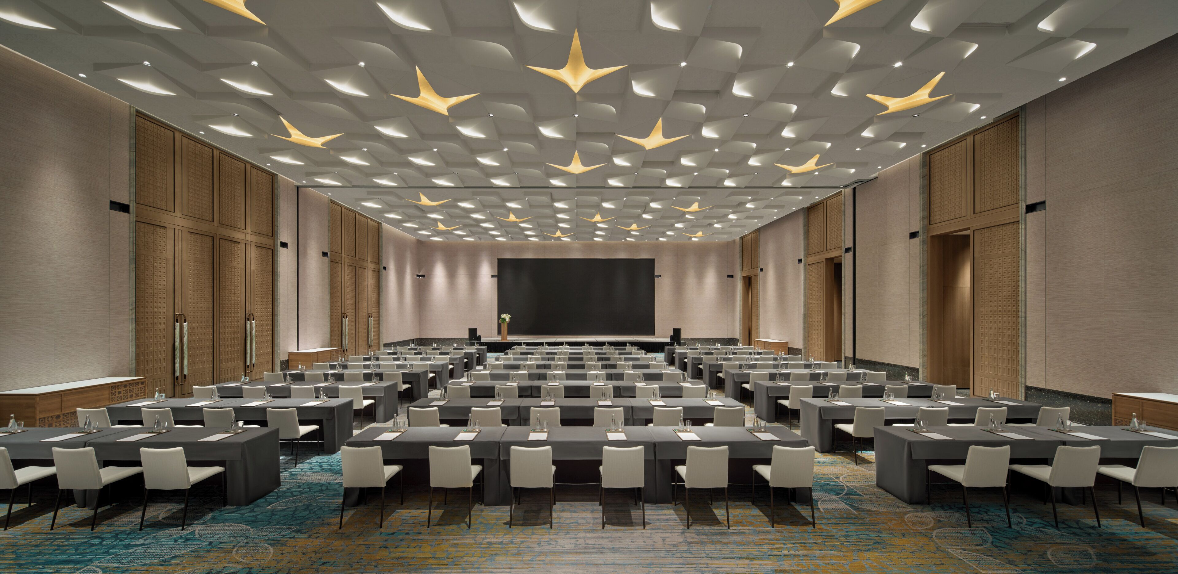 A conference room with rows of tables and chairs, a large screen, and a patterned ceiling.