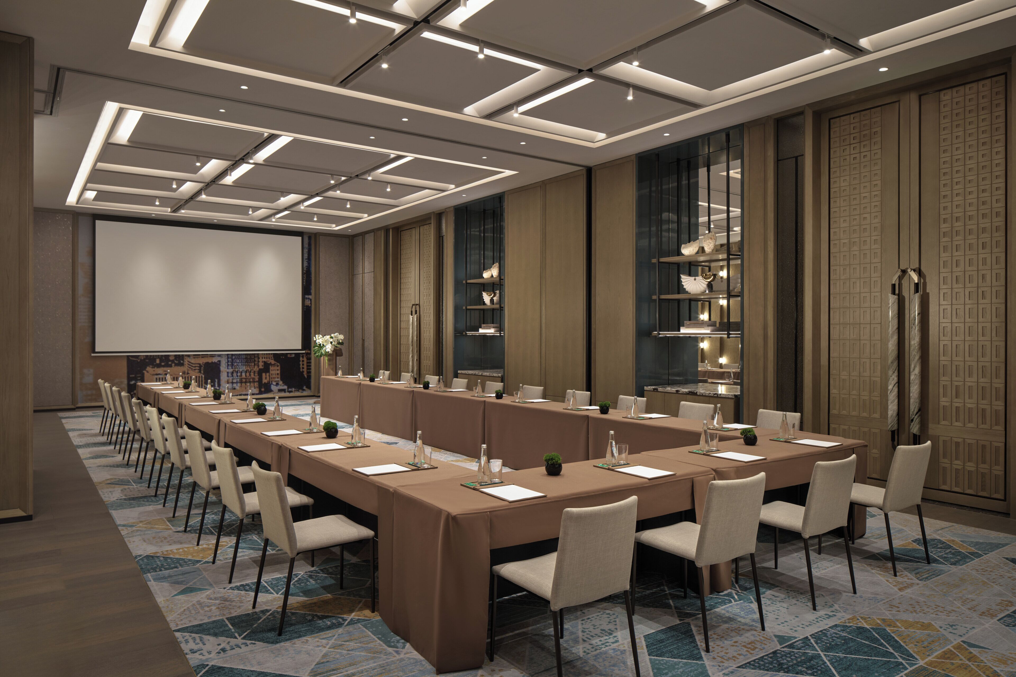 A conference room with a long table, chairs, a projector screen, and a decorative wall with shelves.