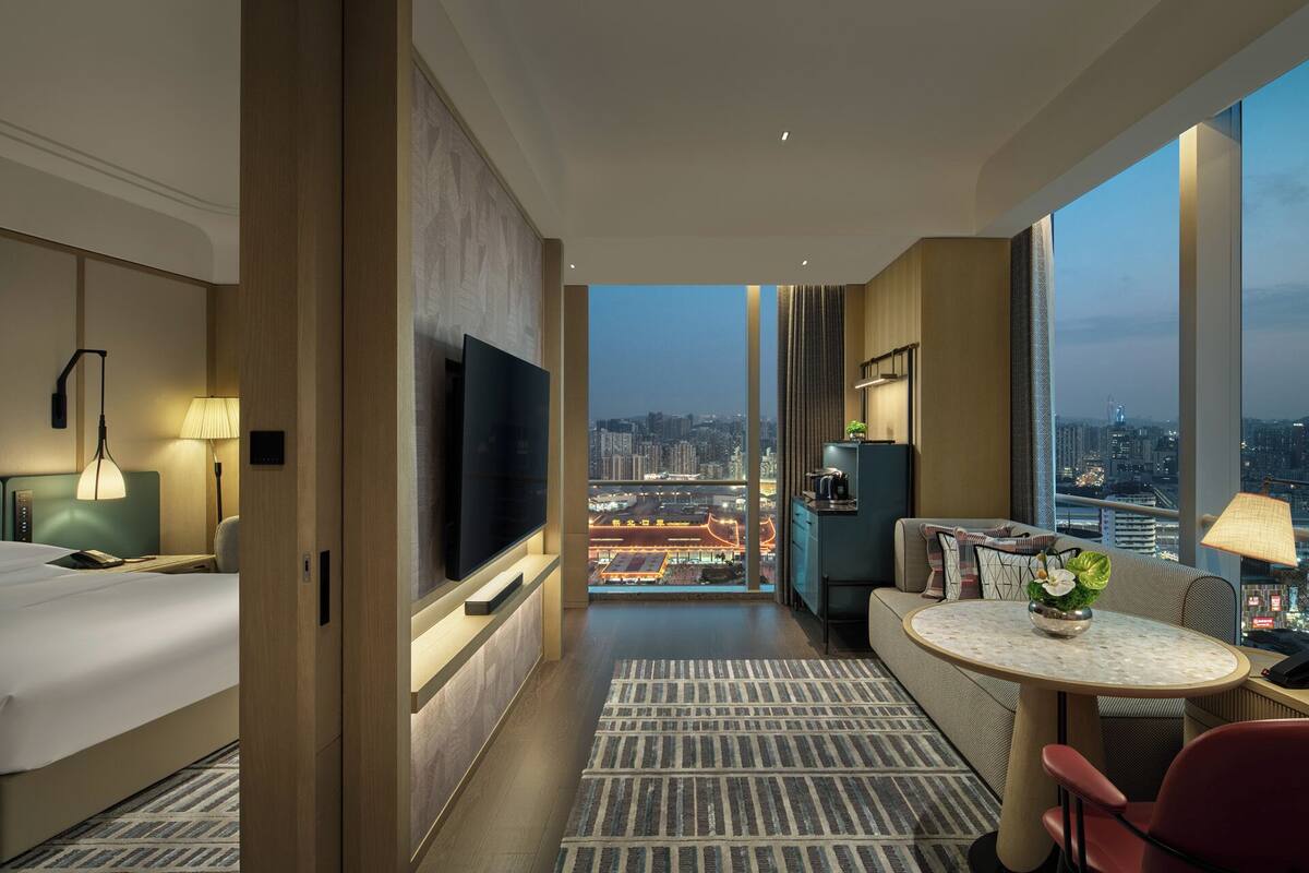 Habitación de hotel con cama, televisor, sofá, una mesita y vista a la ciudad.