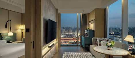 Ein Hotelzimmer mit Bett, Fernseher, Sofa, einem kleinen Tisch und Blick auf die Stadt.