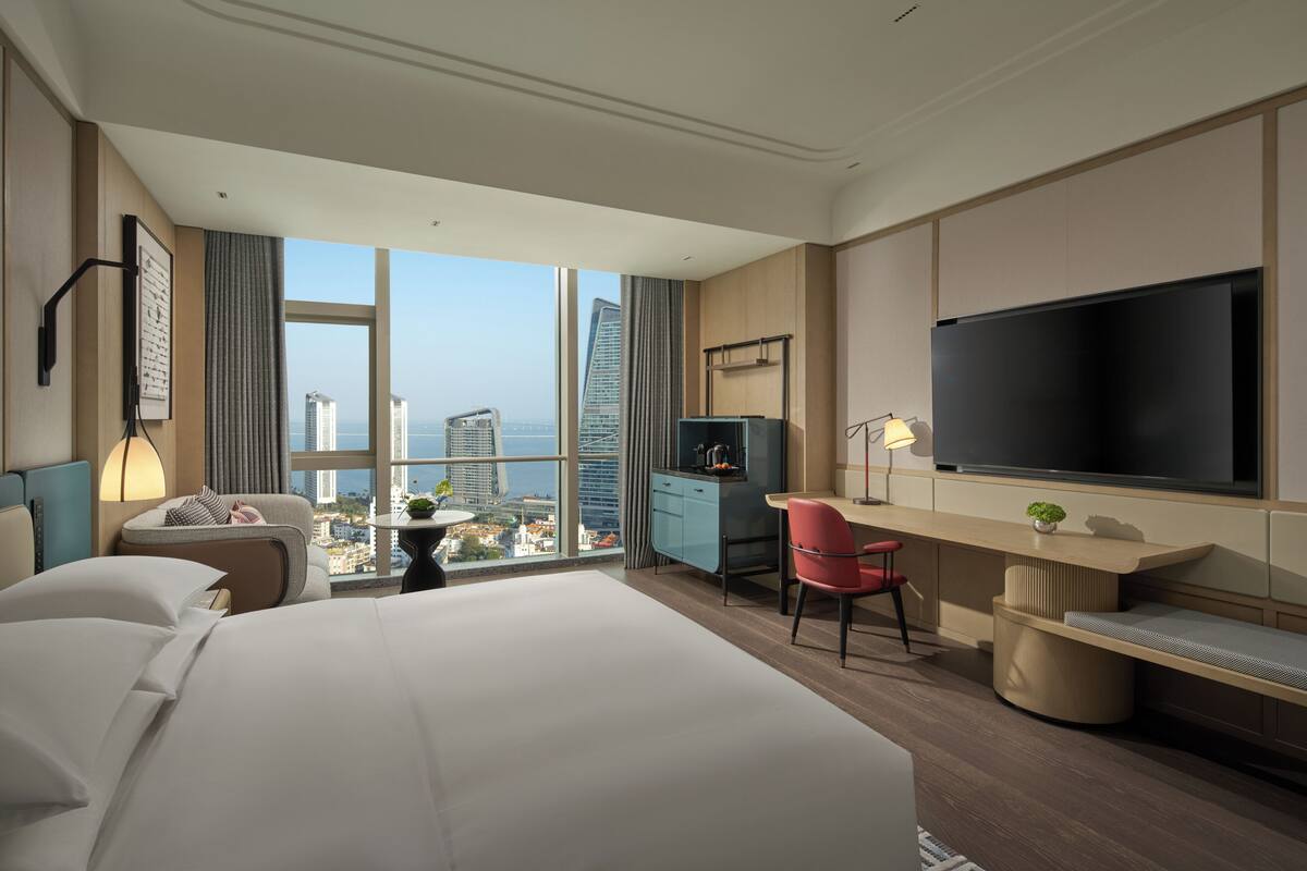 Habitación de hotel con una cama grande, un escritorio con televisor, una silla, una mesita y vistas a la ciudad.