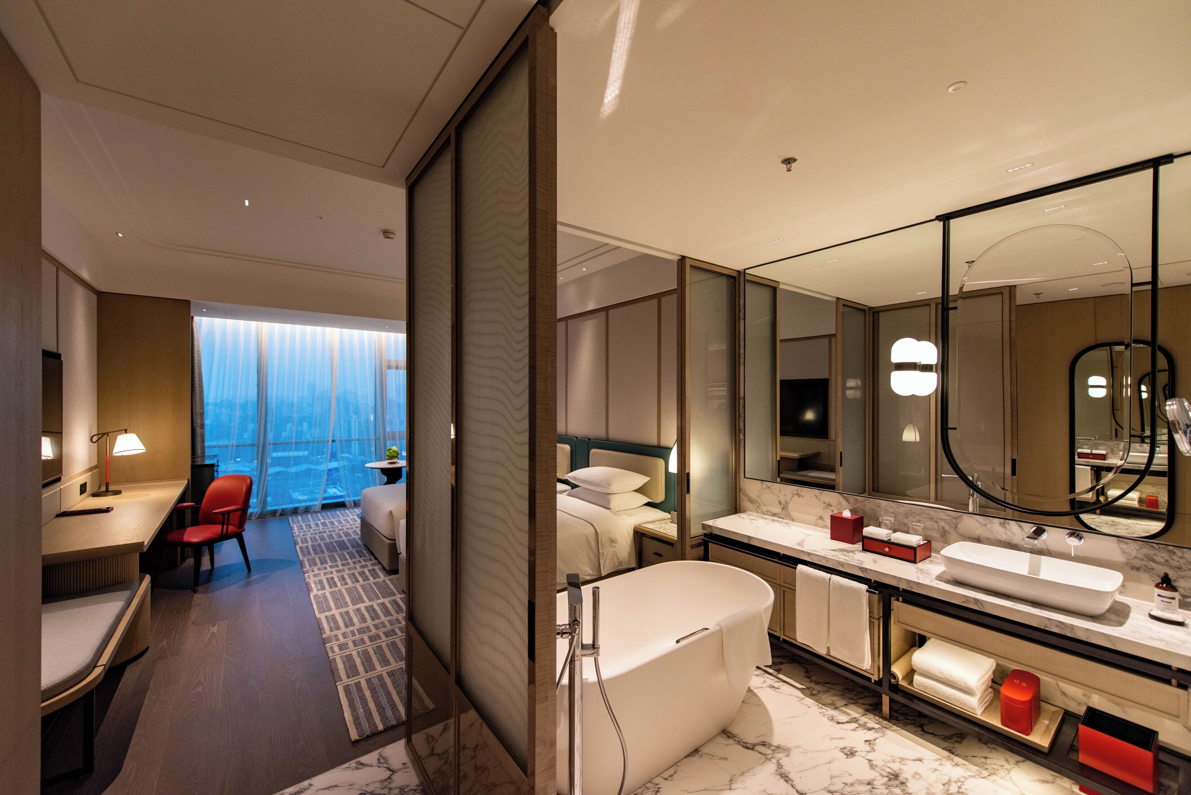 Una habitación de hotel moderna con una cama grande, un escritorio con silla, un baño con bañera y un espejo amplio.