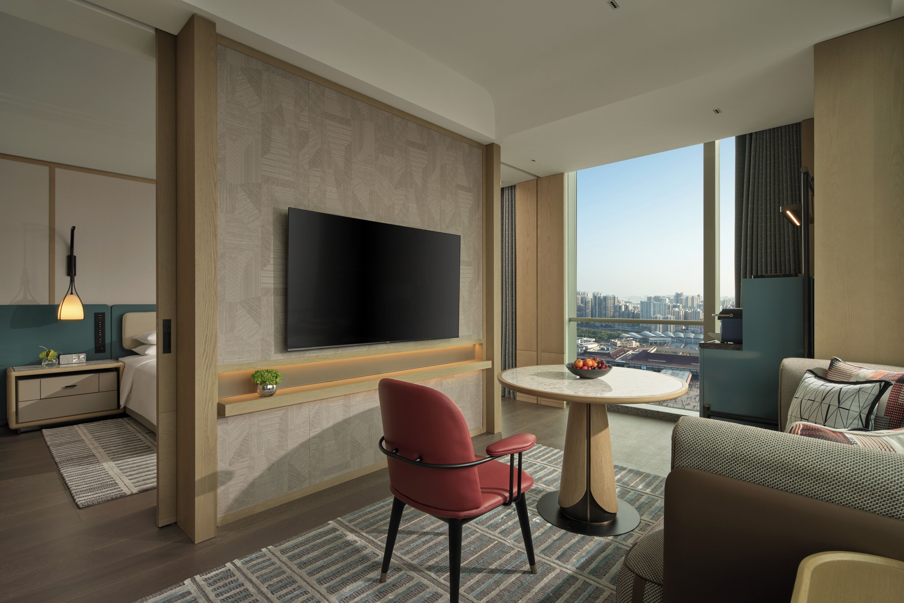 A modern hotel room with a large TV, a dining area with a round table, a red chair, and a city view through large windows.