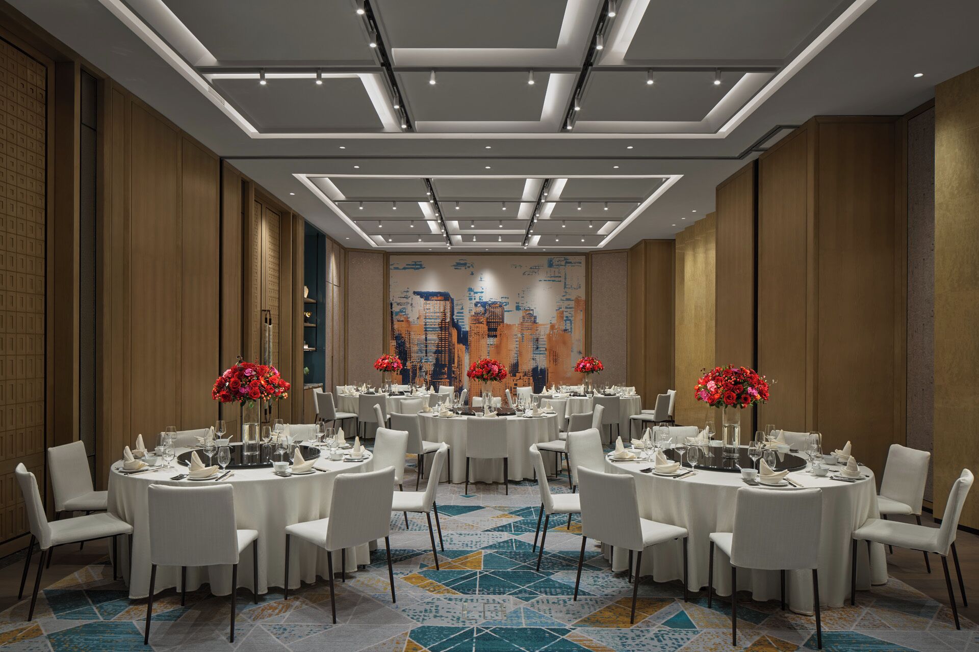 A hotel dining area with round tables, white chairs, and red floral centerpieces.