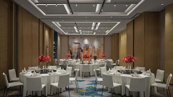A hotel dining area with round tables, white chairs, and red floral centerpieces.